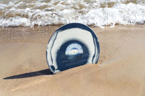 PSD mockup of a colored vinyl record featuring a swirling blue and white design placed on sandy beach by the water.
