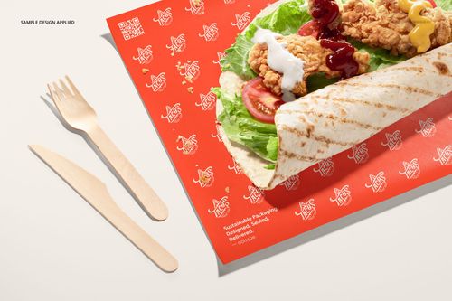 Mockup scene showing a tortilla wrap on bright orange paper with white logo prints, next to wooden fork and knife.