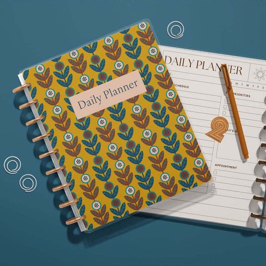 Top view of two spiral planners on a blue surface, one with a floral patterned cover and one open to a daily planner page.