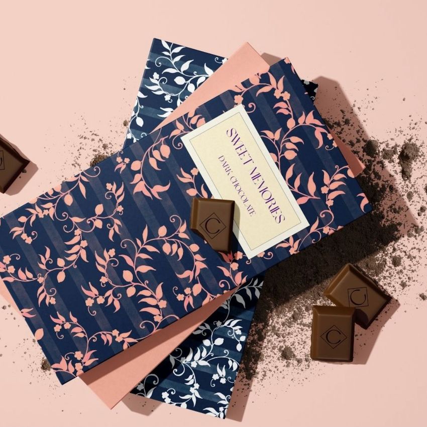 Chocolate bar packaging mockup featuring a navy blue wrapper with pink floral patterns, chocolate pieces, and cocoa powder on a light pink surface.