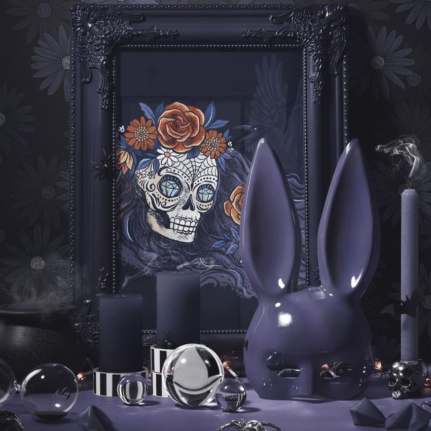 Halloween art print mockup featuring an ornate dark picture frame with a skull illustration, surrounded by purple candles, a bunny mask, and glass spheres on a patterned surface.