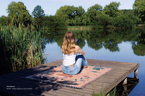 Jacquard fringed throw blanket mockup displayed outdoors on a dock, with a person seated near water and lush trees.