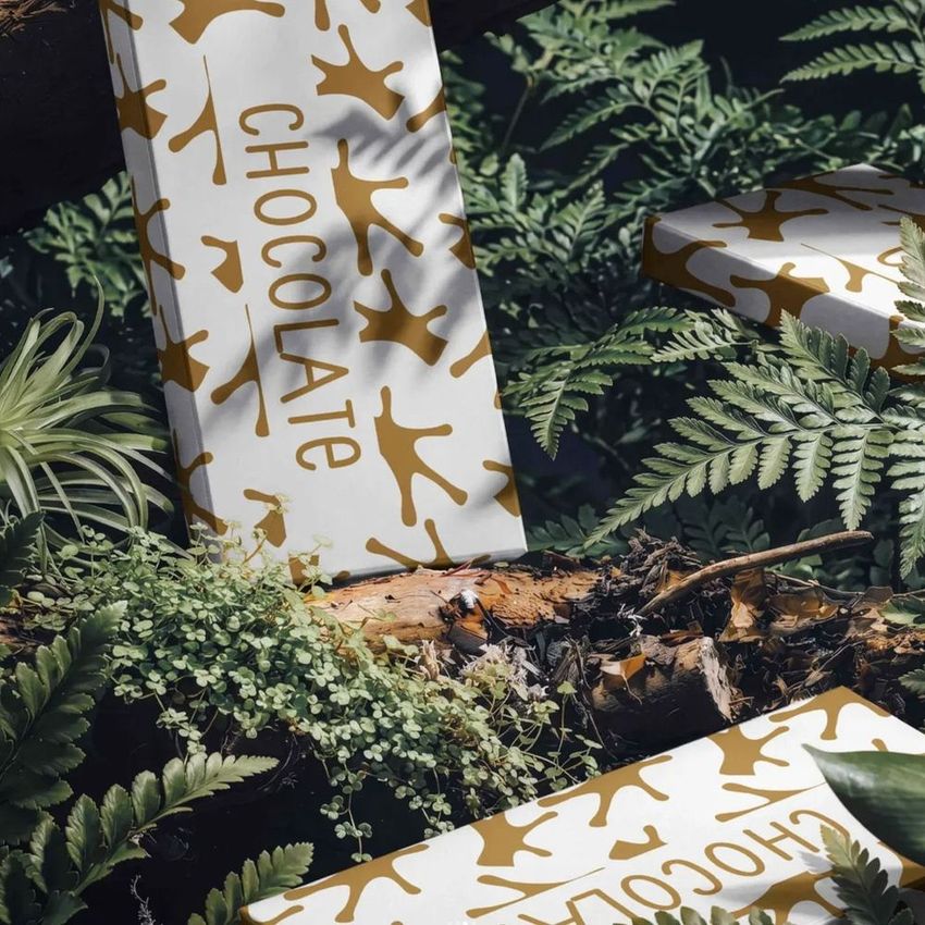 Chocolate box mockup featuring white packaging with gold abstract patterns, placed among green ferns and moss in a forest setting.