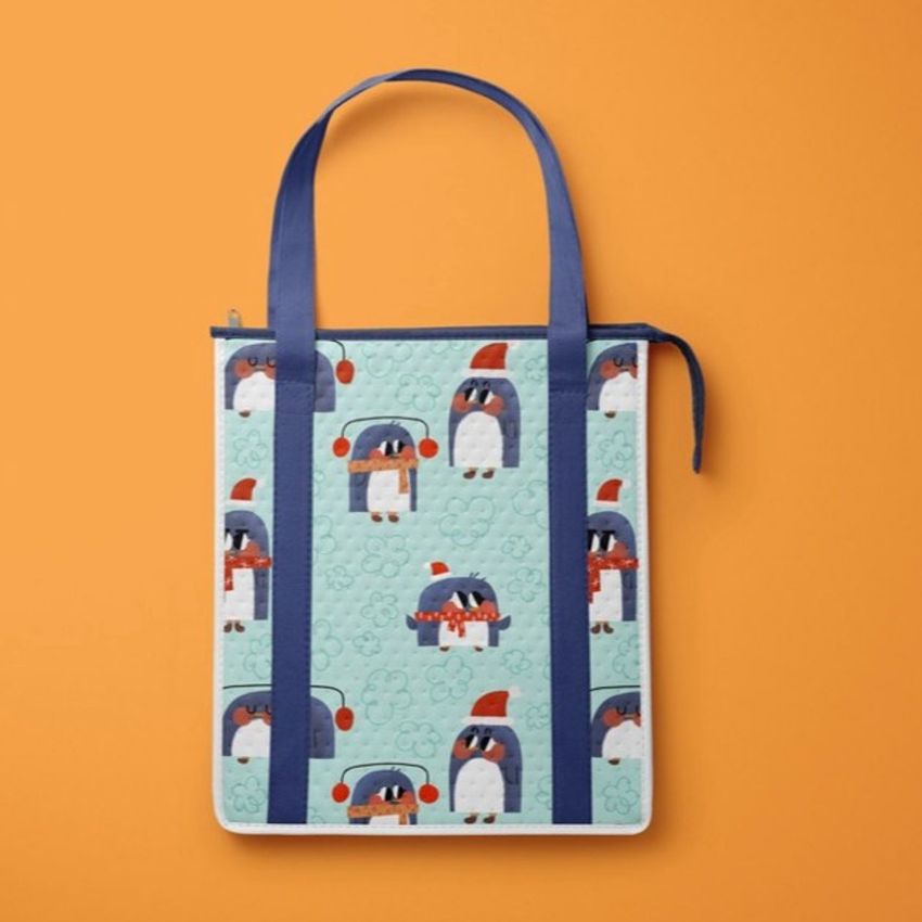 Insulated cooler bag mockup featuring a playful penguin pattern with blue handles, set against a solid orange background.