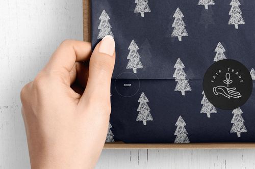 Close-up of a hand lifting navy tissue paper with white tree illustrations inside a cardboard mailer box mockup.