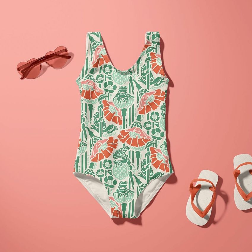 Baby swimsuit mockup displayed on a pink background with a green and orange frog pattern, sunglasses, and flip-flops nearby.