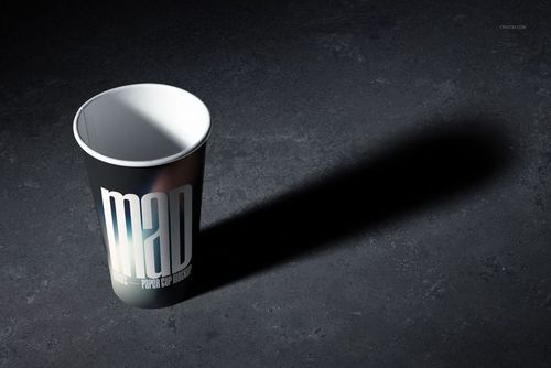 PSD mockup featuring a paper cup with a dark design and prominent text, illuminated and casting a shadow on stone-like ground.