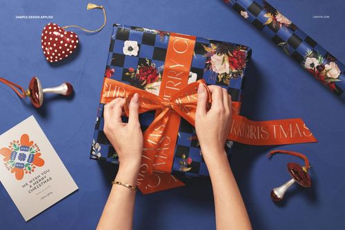 Christmas gift mockup featuring hands arranging a ribbon on a patterned box, with wrapping paper and ornaments nearby.