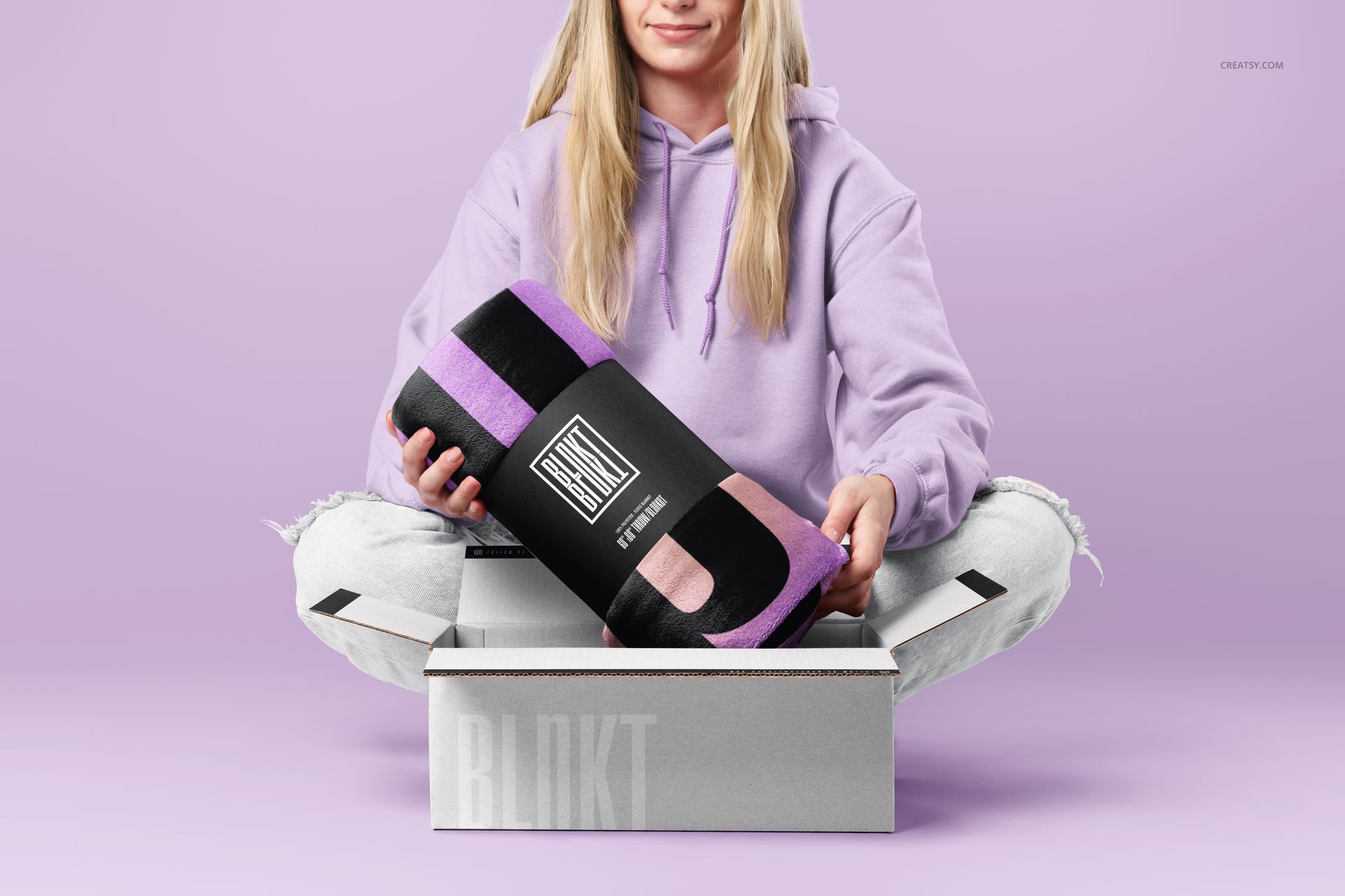 Front view of a fleece blanket mockup, rolled and labeled, presented by a woman seated on a light purple background.