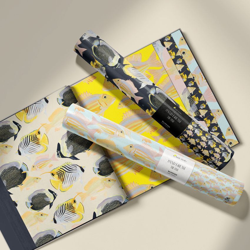 Wallpaper sample book mockup featuring open pages with colorful fish patterns and two rolled wallpaper samples on top.