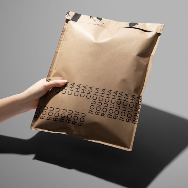 Hand holding a kraft bubble mailer mockup with repeating black text pattern on a neutral gray background, casting a shadow.