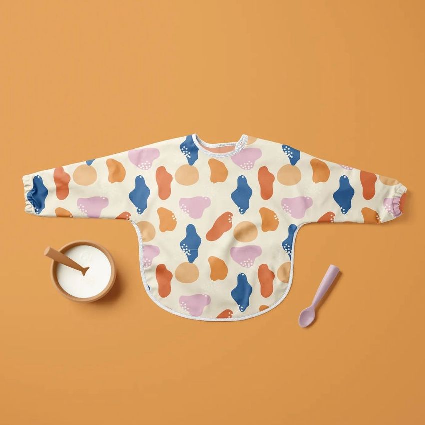 Baby sleeved bib mockup with colorful abstract shapes in blue, orange, and pink on a beige background, shown on a solid orange surface.