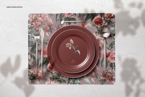 Fringed placemat mockup shown from above with decorative floral print, maroon plates, and metallic utensils on white surface.
