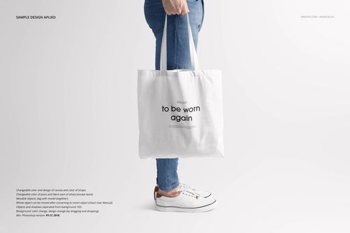 Front view of a white tote bag mockup held by a person wearing blue jeans and white sneakers against a plain background.