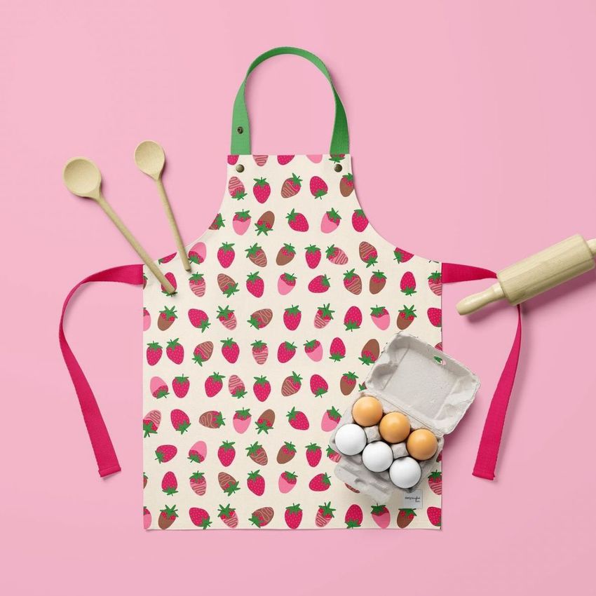 Kids apron mockup featuring a strawberry pattern, displayed on a pink background with wooden spoons, eggs, and a rolling pin.
