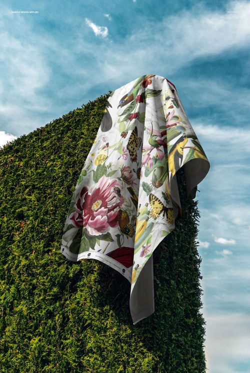 PSD mockup of a fabric featuring vivid floral and wildlife design, displayed outdoors on a hedge with a blue sky backdrop.