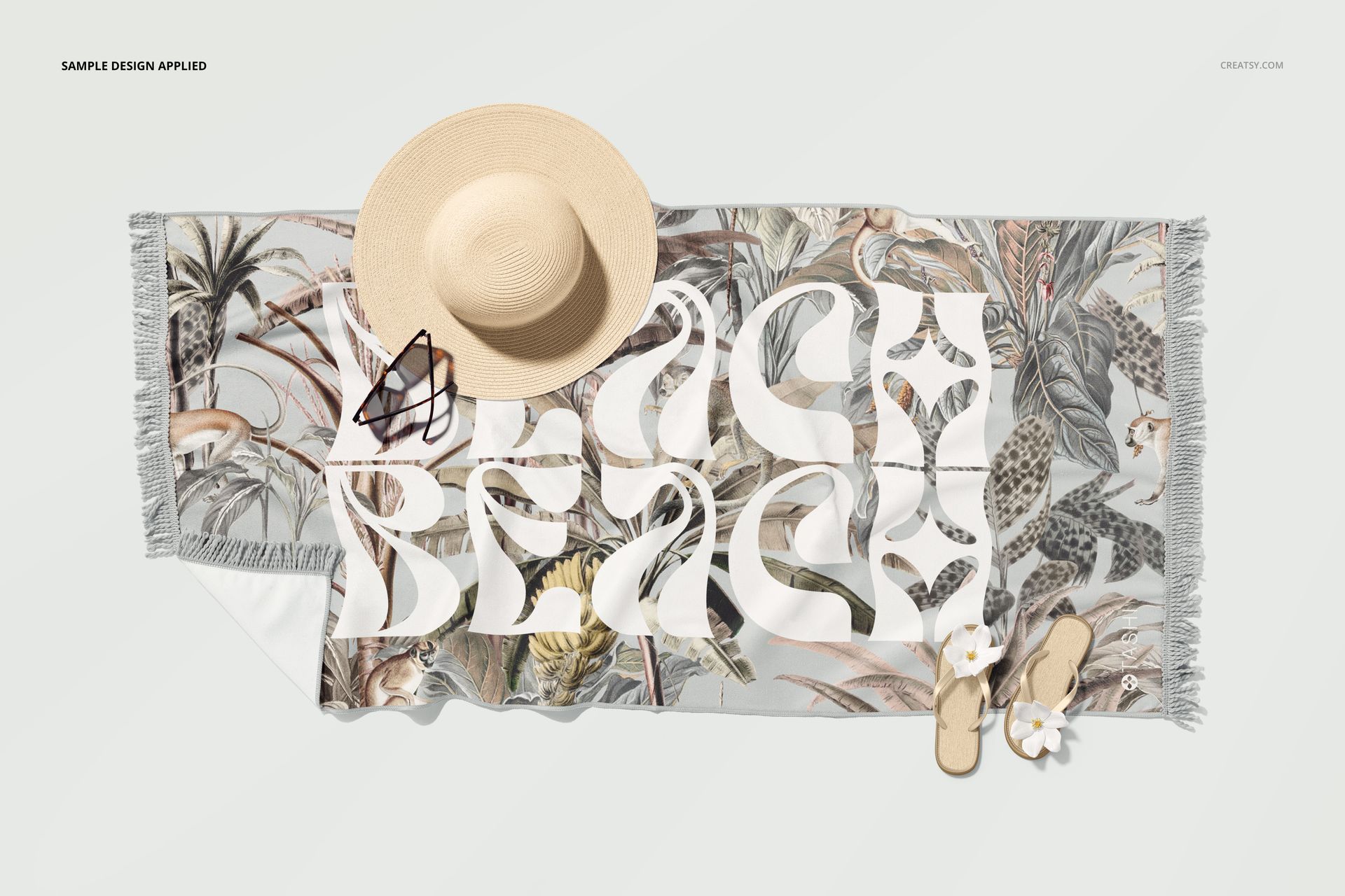 Top view of a fringed beach towel mockup featuring a jungle motif, surrounded by a sun hat, sandals, and flowers.