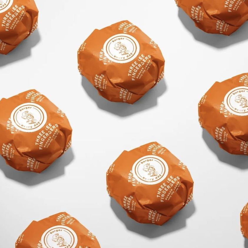 Burger mockup featuring several burgers wrapped in orange paper with a white circular logo and repeating text, arranged on a light surface.