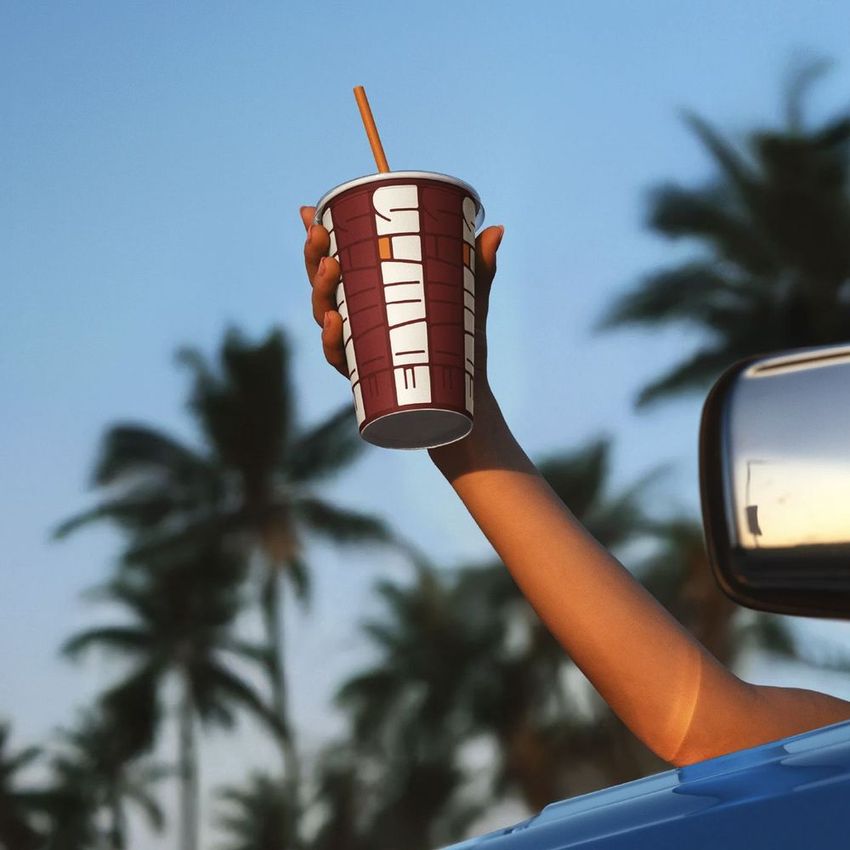Hand holding a brown beverage paper cup mockup out of a car window, with palm trees and blue sky in the background.