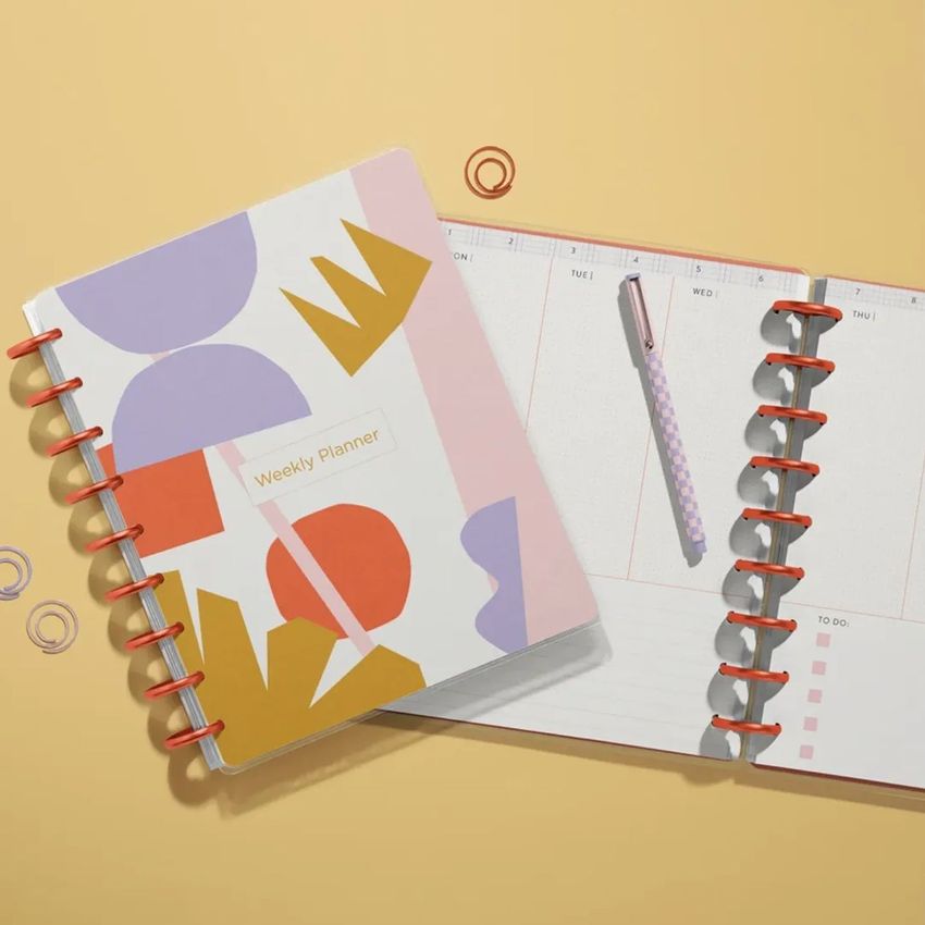 Two spiral-bound planner mockups seen from above, featuring pastel abstract shapes and a pen on a light tan background.