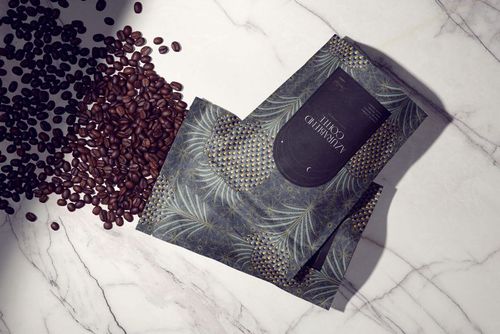 Coffee packaging mockup with dark patterned bags and scattered coffee beans on a white marble surface, top view.