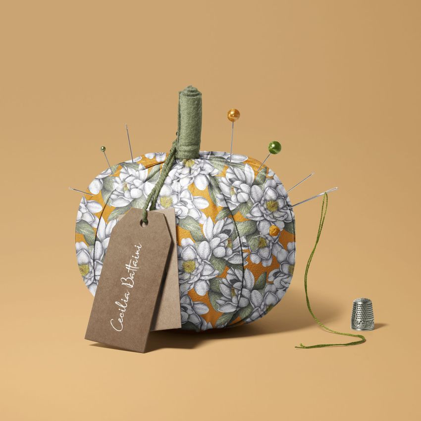 Fabric pumpkin mockup with a white and green floral pattern, sewing pins, a brown tag, and a thimble on a beige background.