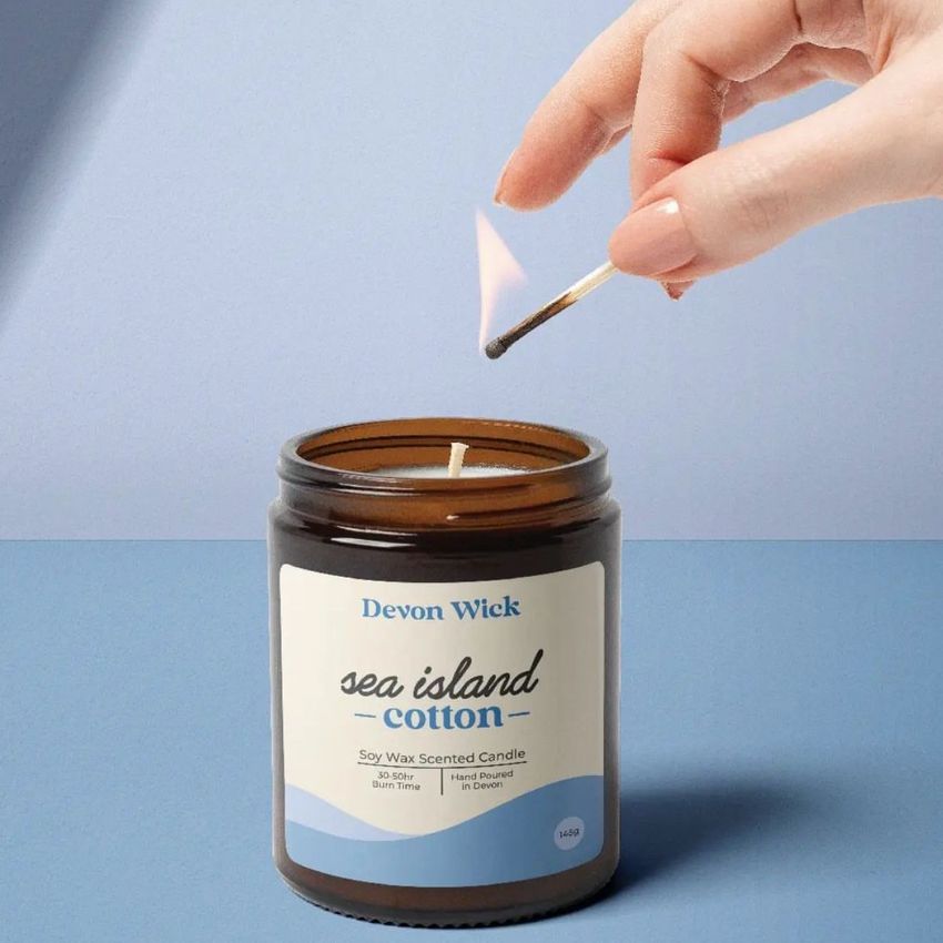 Amber jar candle mockup with a blue and white label on a flat surface, a hand lighting the wick with a match above the candle.