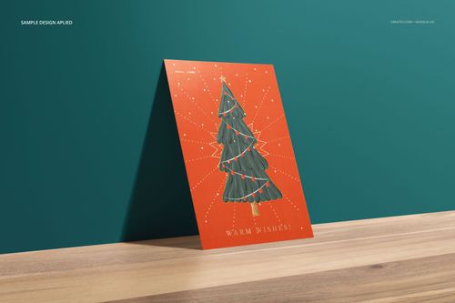Stationery card mockup featuring a vertical card with a green Christmas tree design on an orange background, angled on a wooden surface.