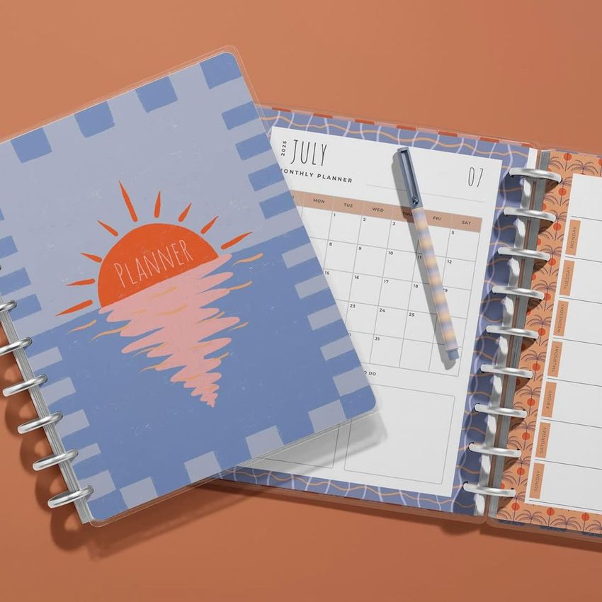 Top view of two spiral planners on a warm orange surface, one closed with a sunset illustration, and one open to a calendar page.