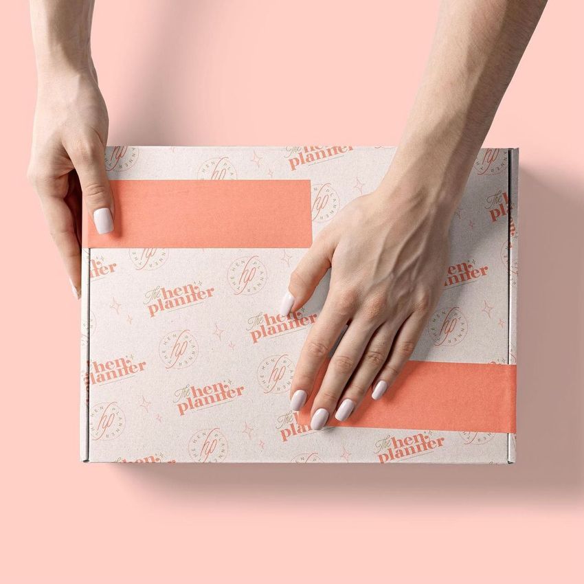 Hands with light nail polish hold a mailing box mockup decorated with orange tape and branded pattern on a pastel pink background.
