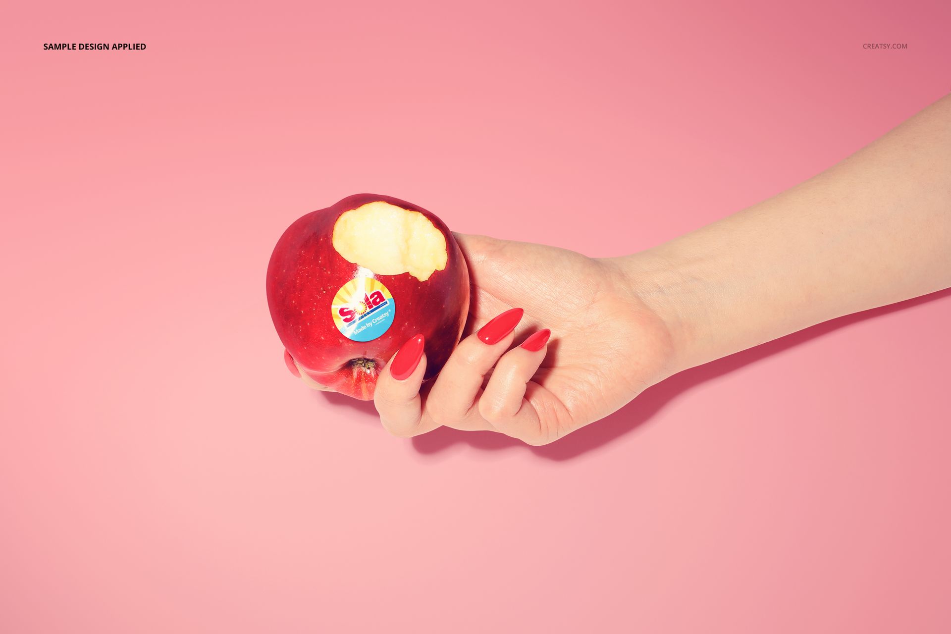 PSD mockup featuring a bitten red apple with a circular sticker, held by a hand with manicured red nails on pink.