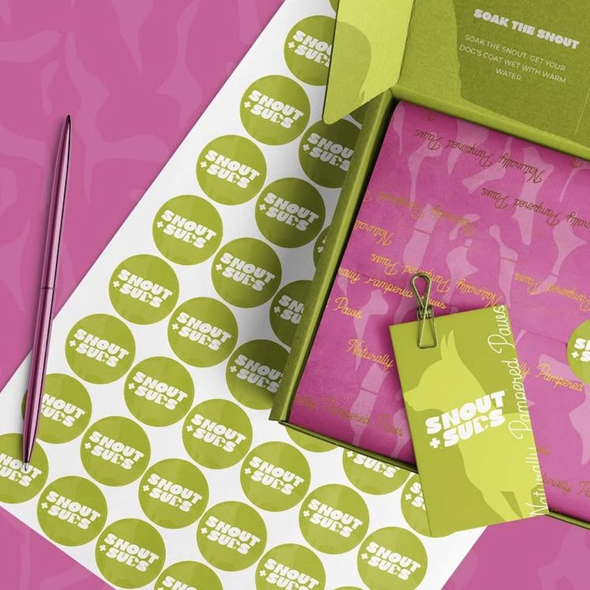 Mailing box mockup featuring an open cardboard box with green and pink branding, stickers, and a pen on a patterned surface.
