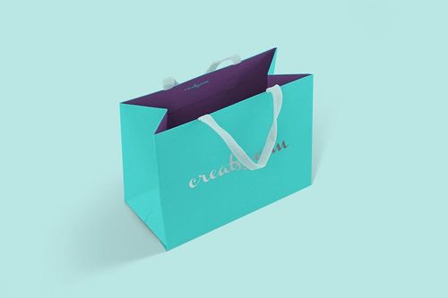 Paper bag mockup featuring a turquoise bag with white rope handles and metallic lettering, angled on a pale blue surface.