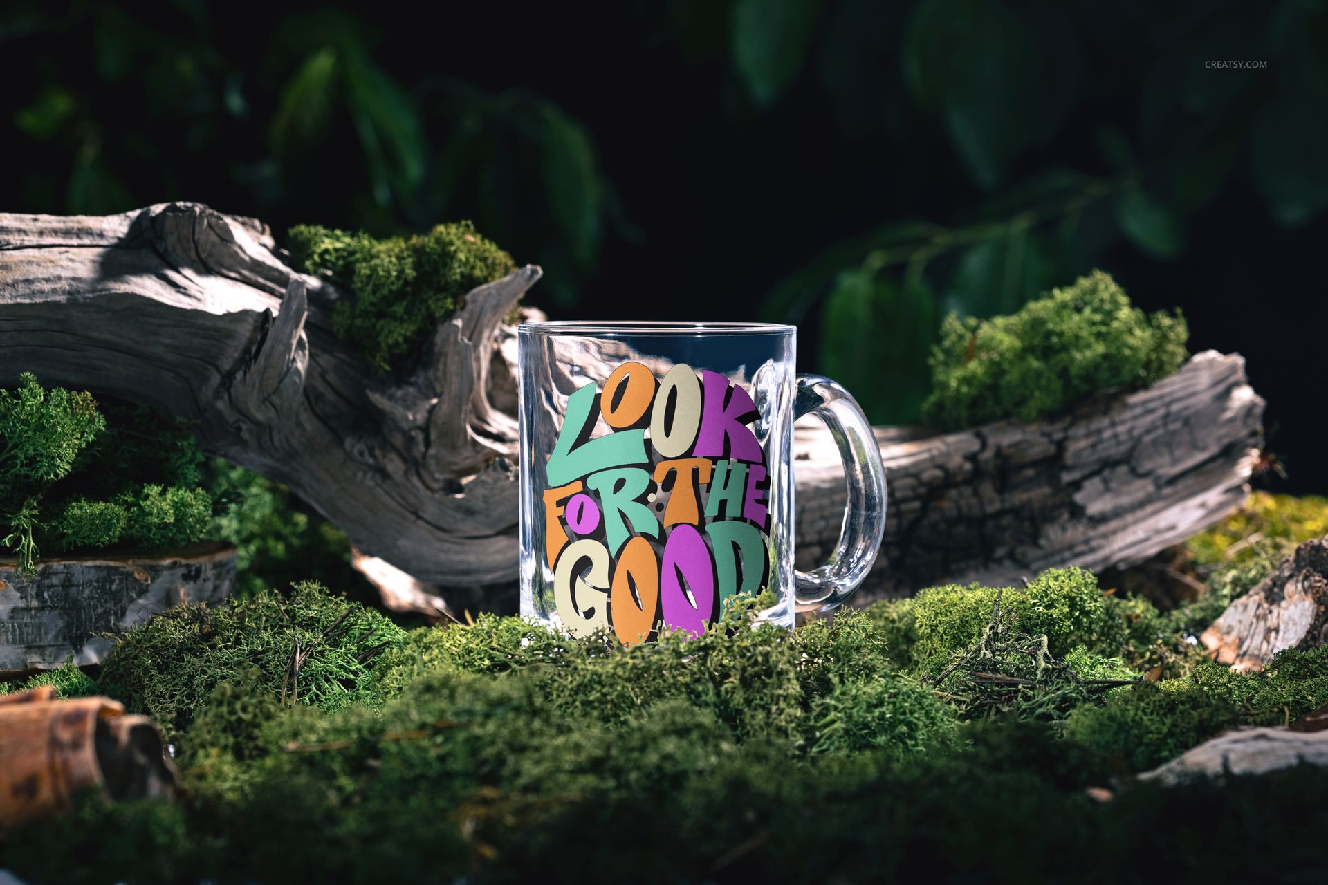 PSD mockup of a clear glass mug displaying vibrant typography, positioned on forest moss among branches and foliage.