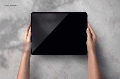 Top view of two hands gripping a dark iPad Pro mockup with an empty display on a textured gray background.