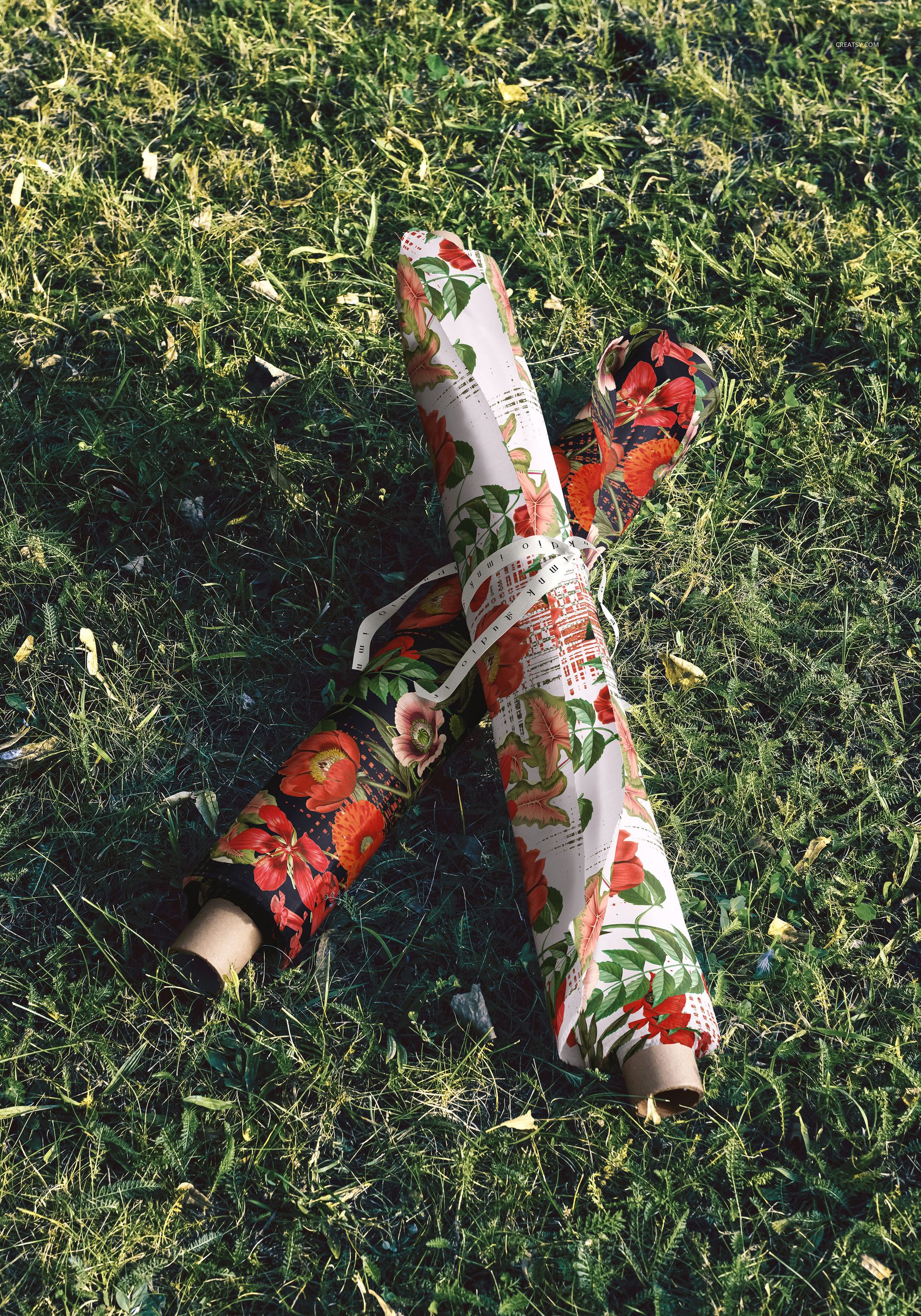 PSD mockup of stacked fabric rolls with red and green floral prints placed outdoors on a grassy surface.