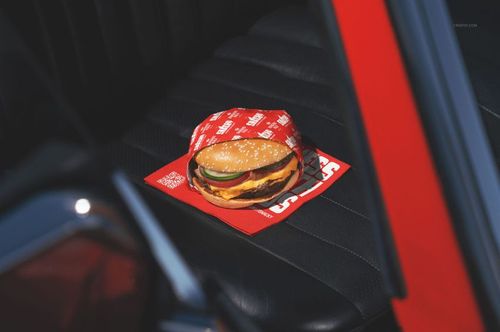Fast food cheeseburger mockup in red packaging, displayed on a dark car interior with sunlight and red trim.
