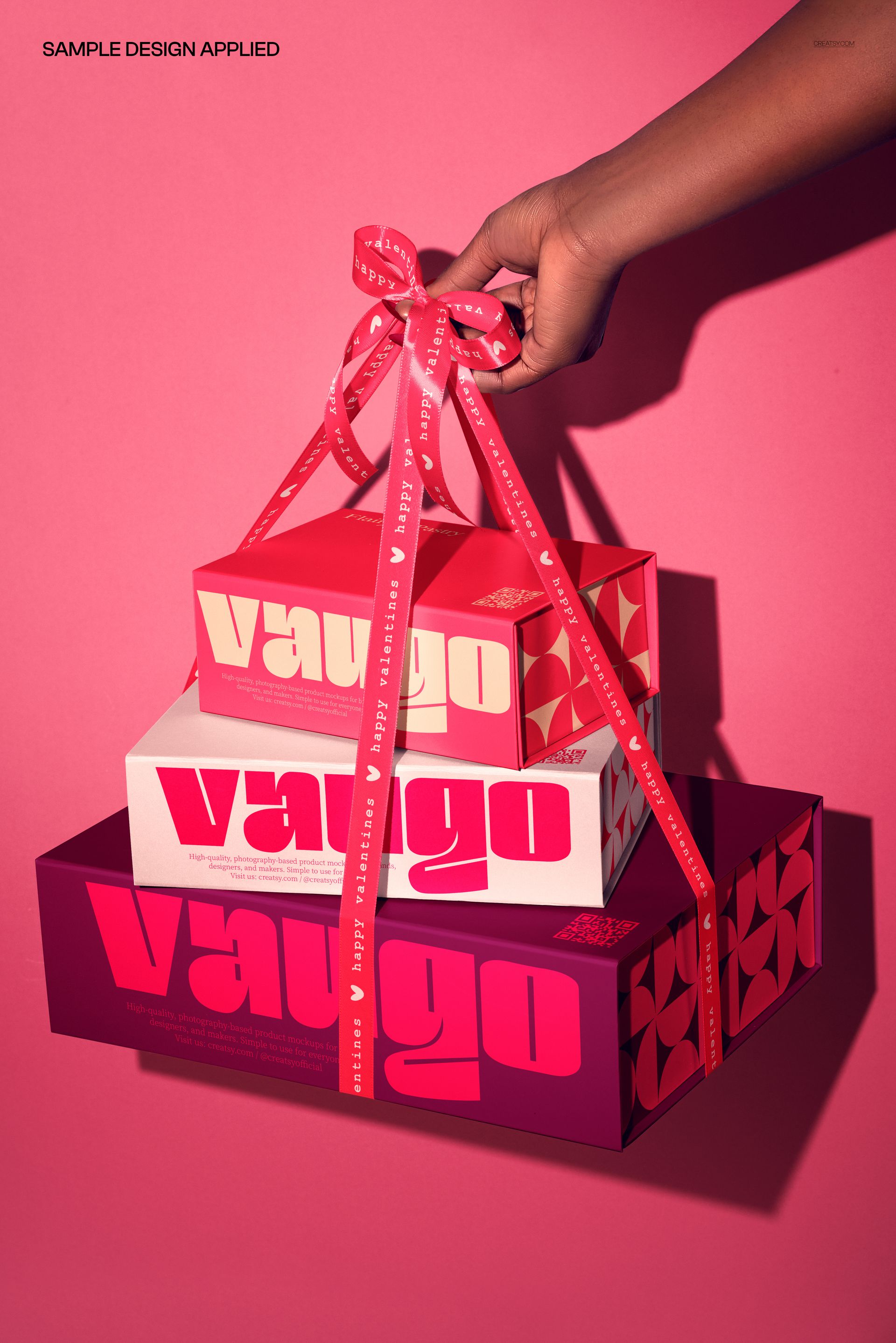 PSD mockup showing a hand gripping ribbon attached to three colorful magnetic boxes, arranged in a stack on a pink surface.