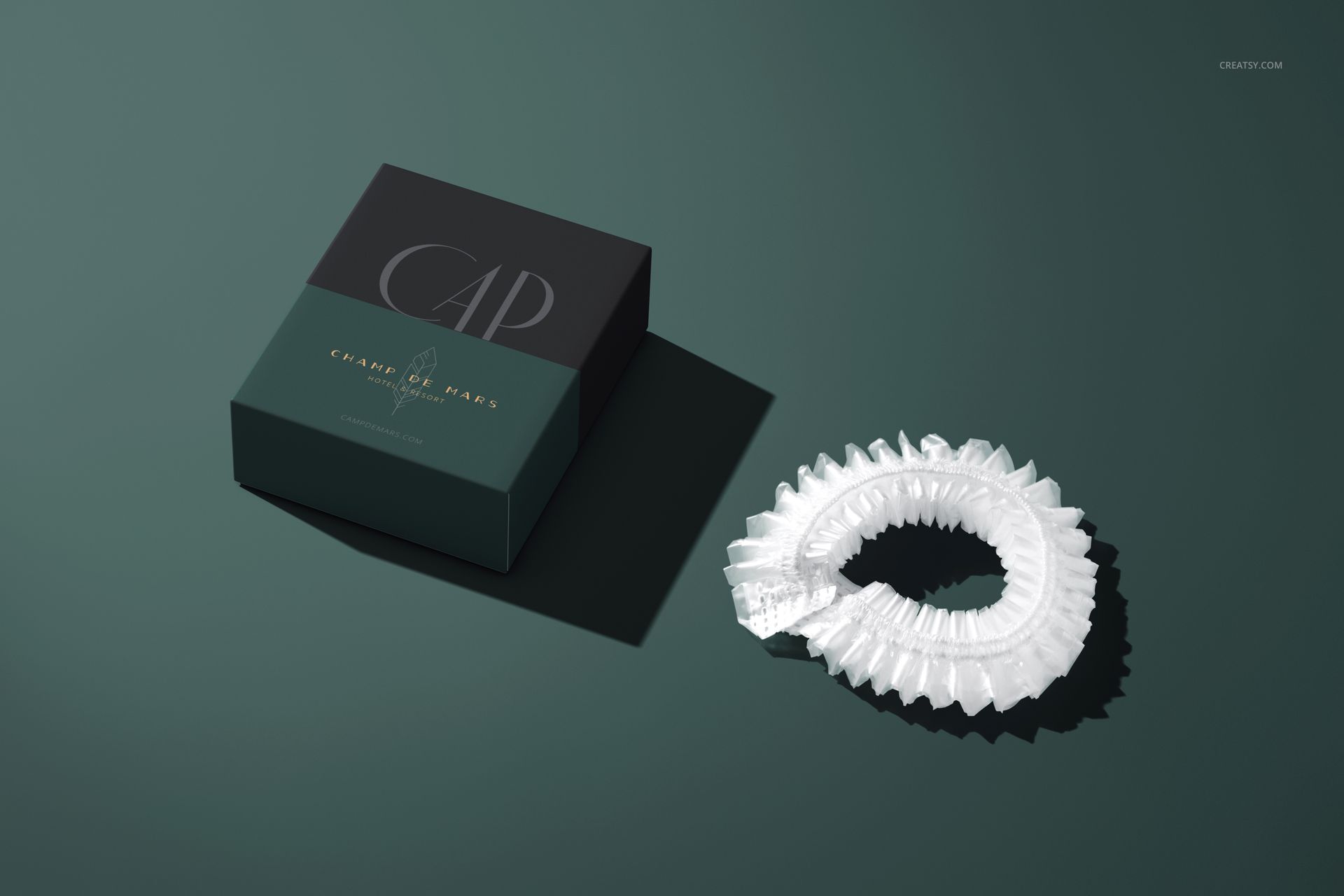 Hotel shower cap mockup showing a clear plastic cap beside a square box, both on a matte green surface.