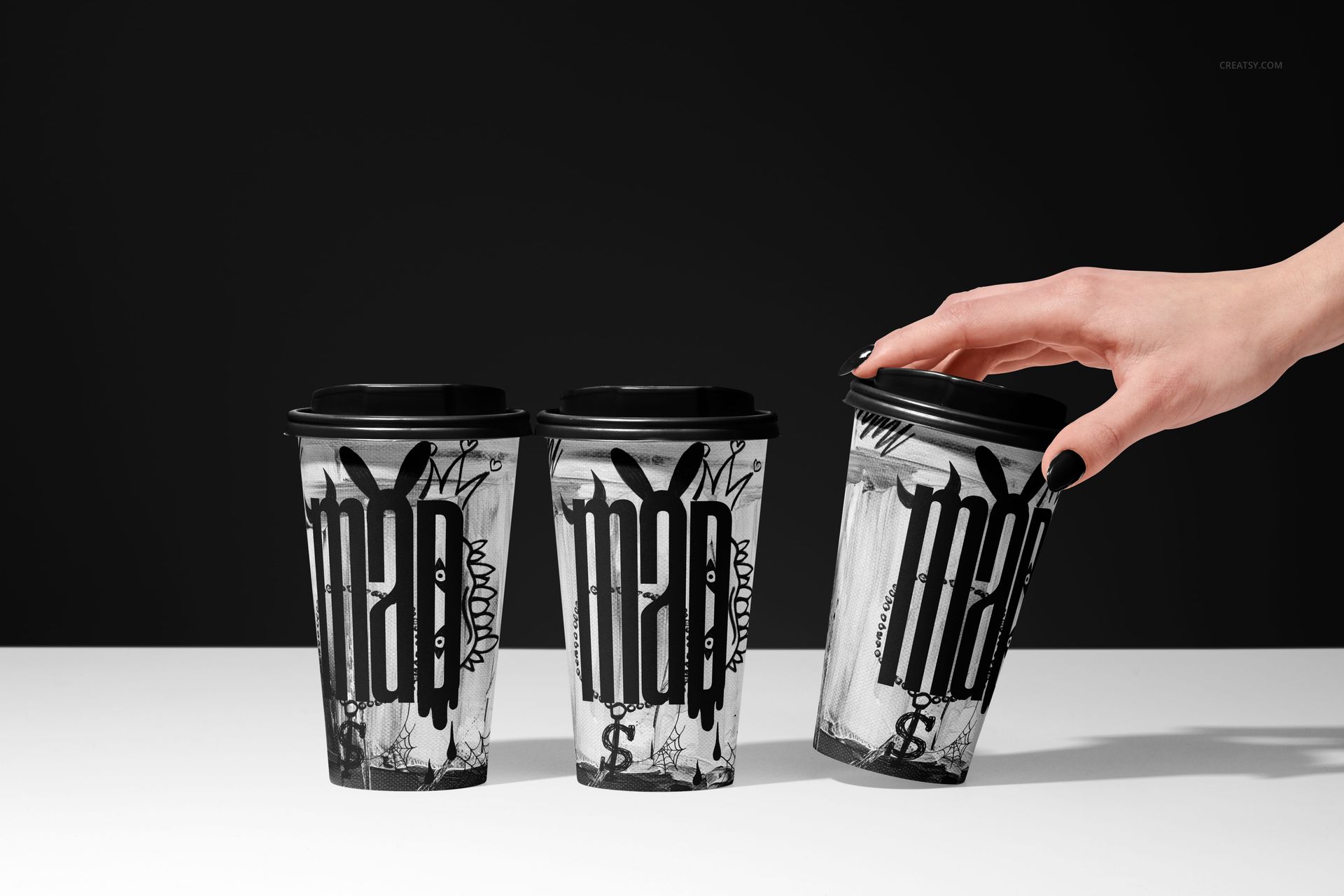 Mockup of three paper coffee cups with bold black and white graphics, black lids, and a hand reaching for one.