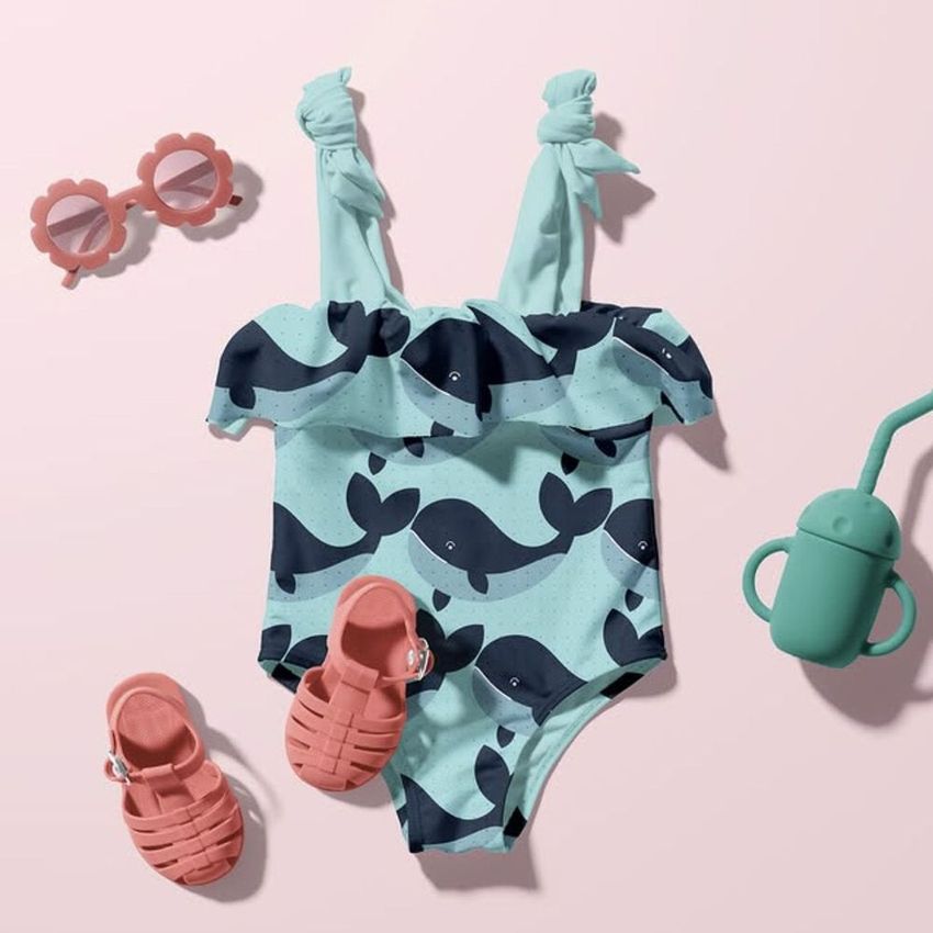 Baby swimsuit mockup featuring a light blue suit with dark whale patterns, displayed flat on a pastel pink surface with accessories.