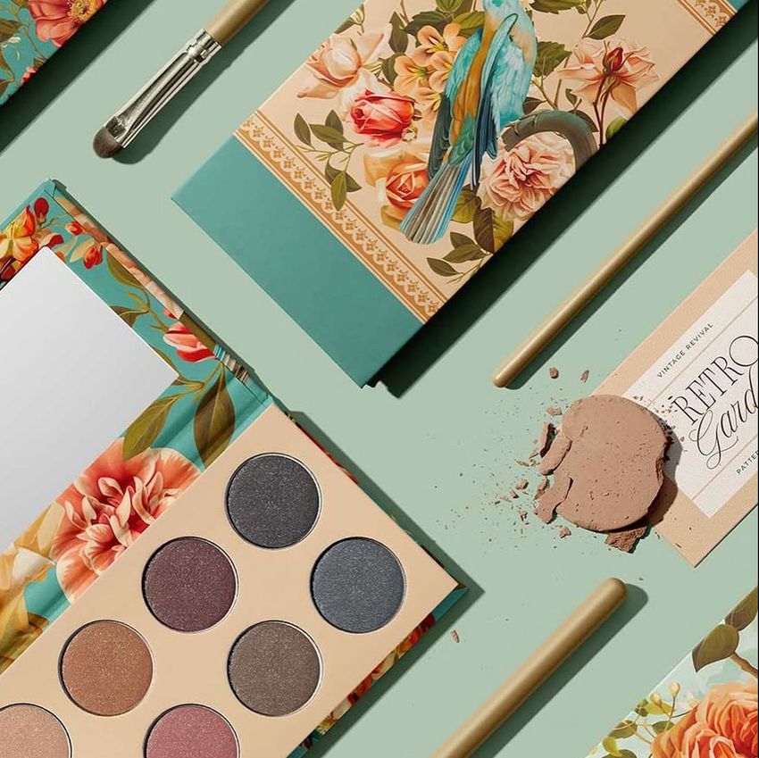 Eyeshadow palette mockup with floral and bird packaging, open to show six round shades, brush, and scattered powder on a green surface.
