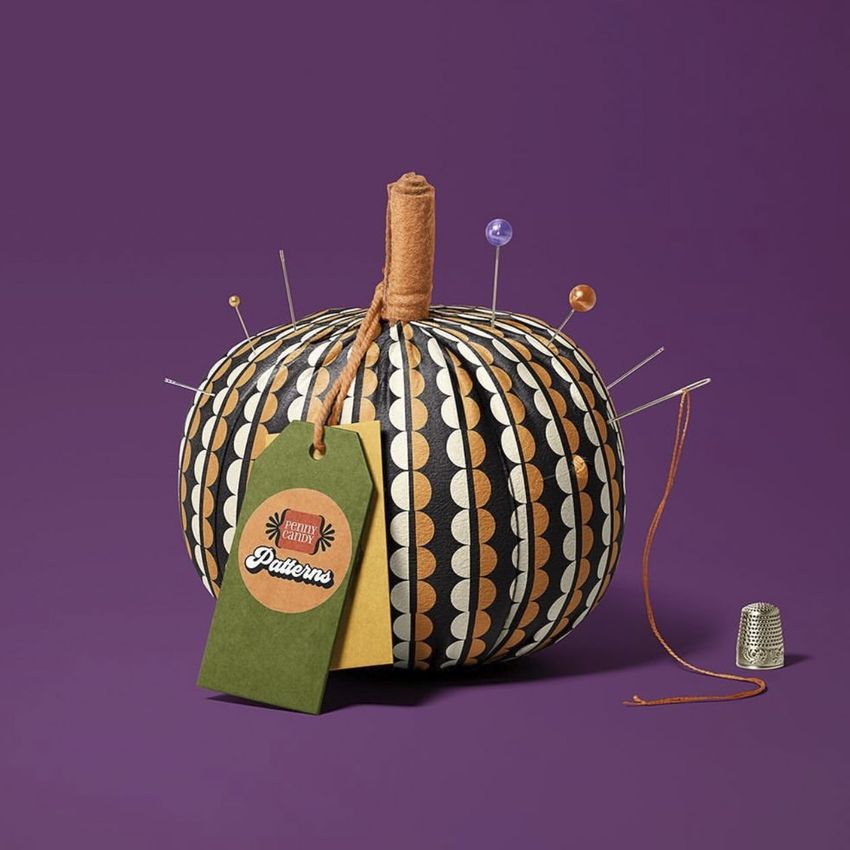 Fabric pumpkin mockup with a geometric black and cream pattern, pins inserted on top, and a green tag, set against a purple background.