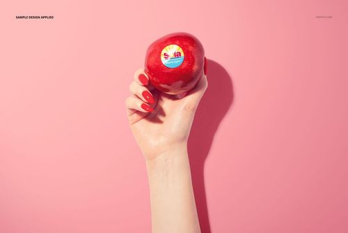 Hand holding a red apple with a round fruit sticker mockup on a solid pink background, front view.