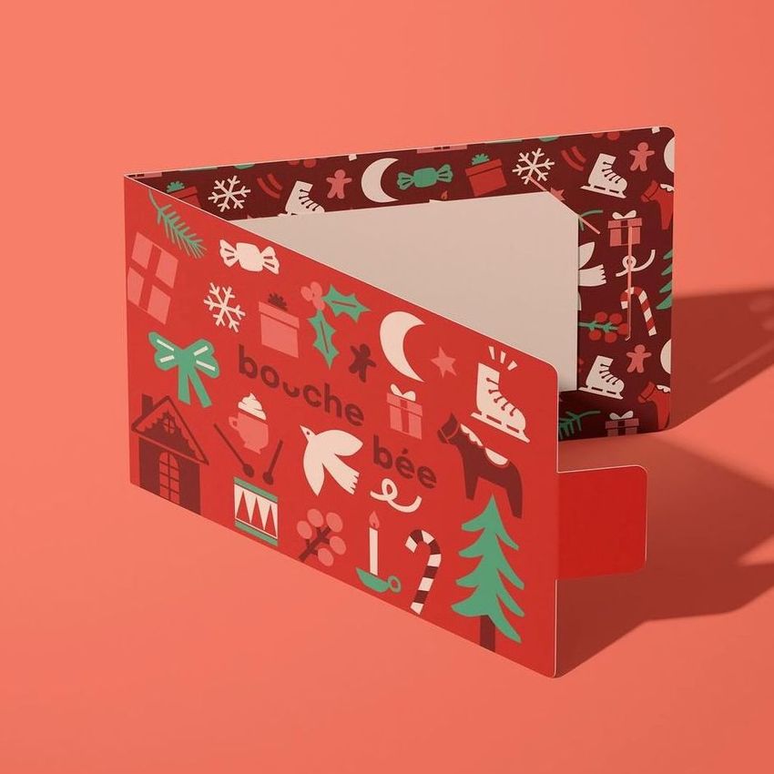 Gift card holder mockup featuring a folded card with festive holiday illustrations in red, green, and white on a coral background.