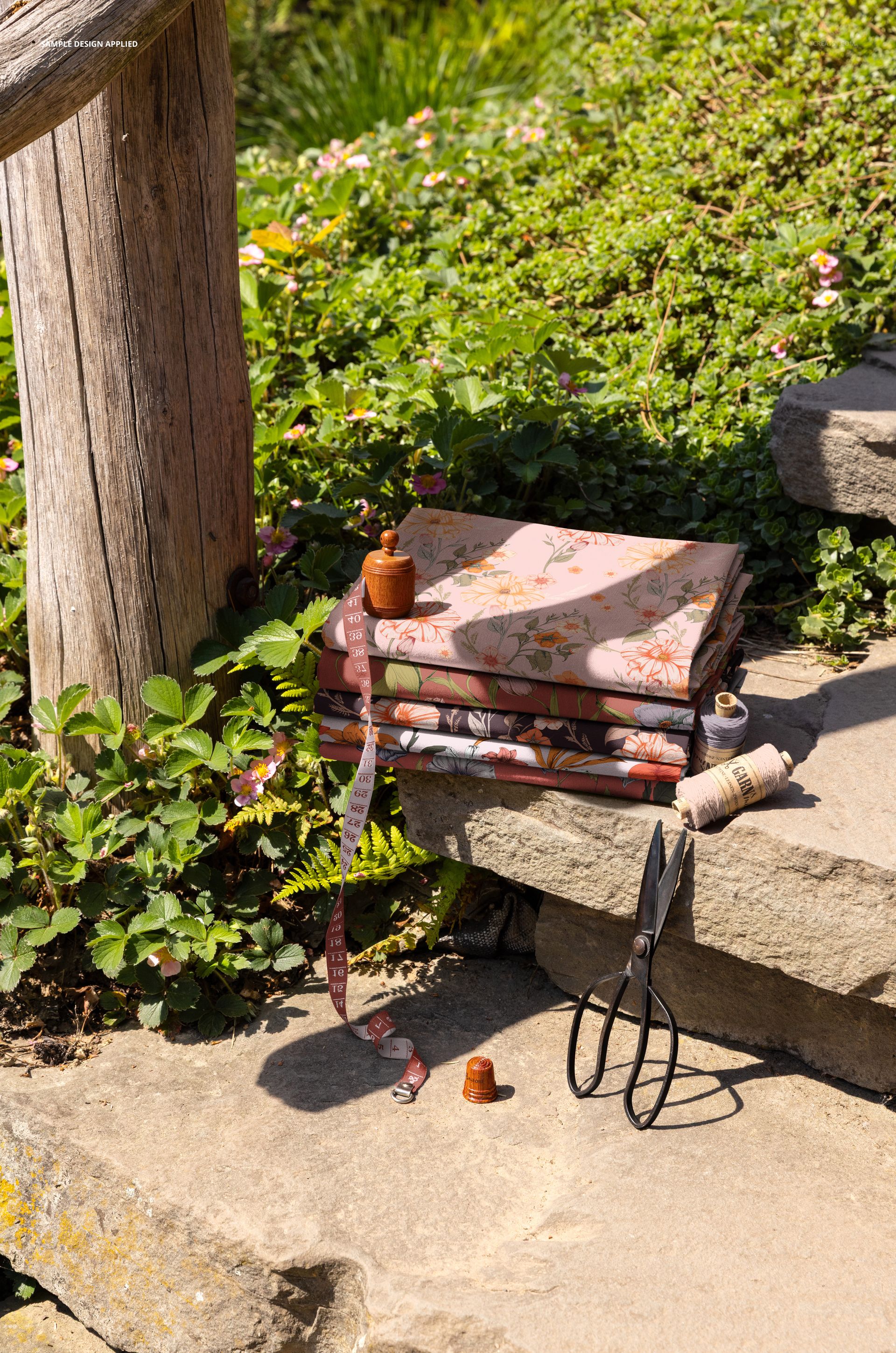 PSD mockup showing a pile of patterned fabrics on rustic stone steps in a garden, accompanied by sewing accessories and lush foliage.