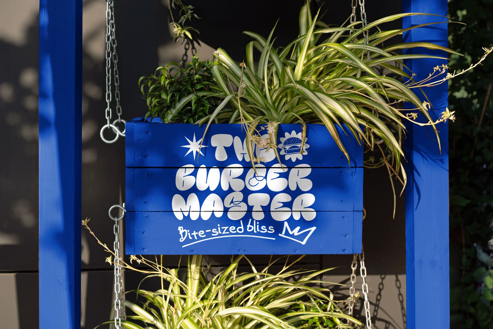 Front view of a blue wooden sign with bold white text and playful graphics, surrounded by green leafy plants.