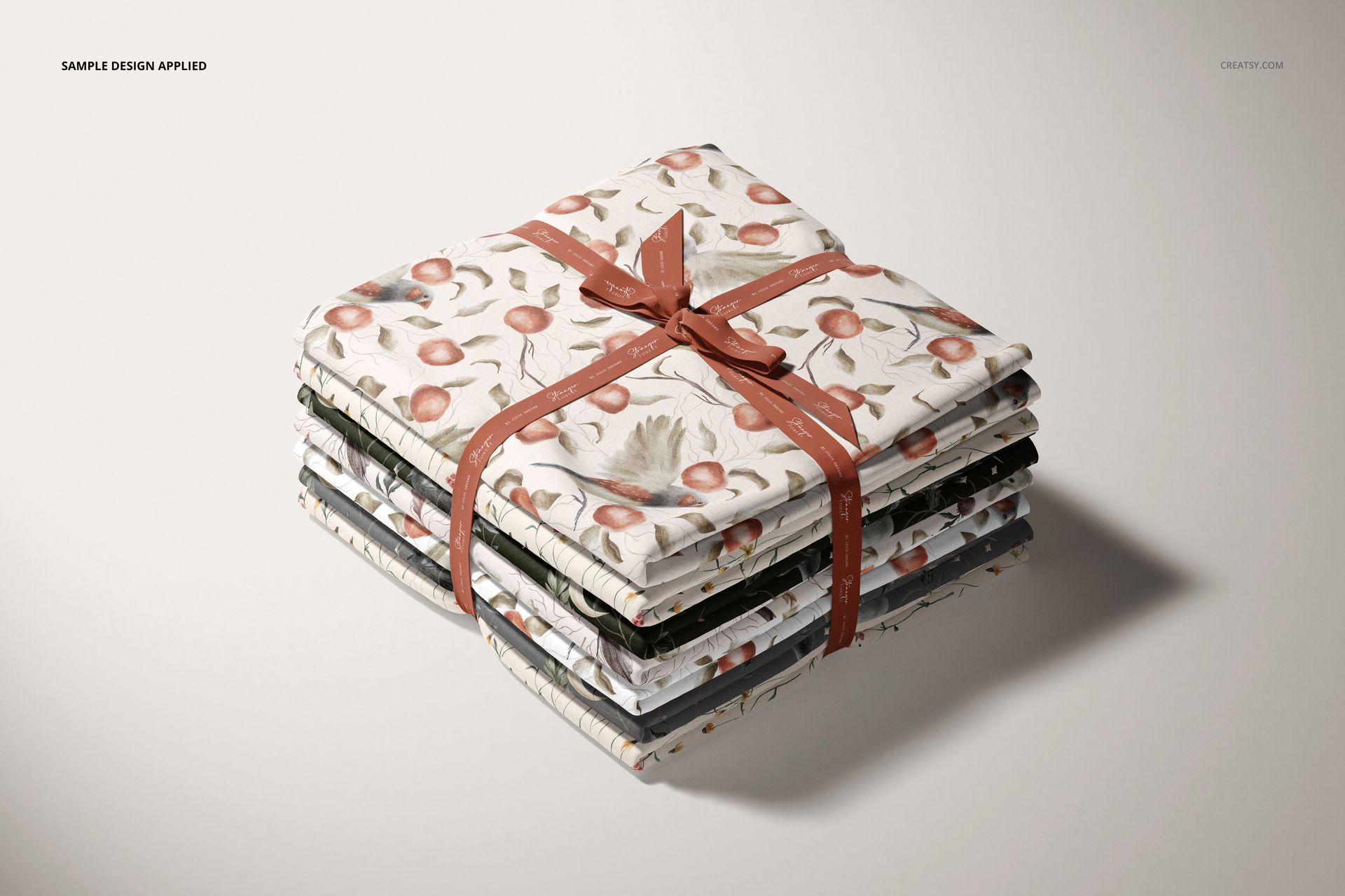 Fabric stack mockup showing multiple folded textiles with nature-inspired prints, tied together with a reddish ribbon.