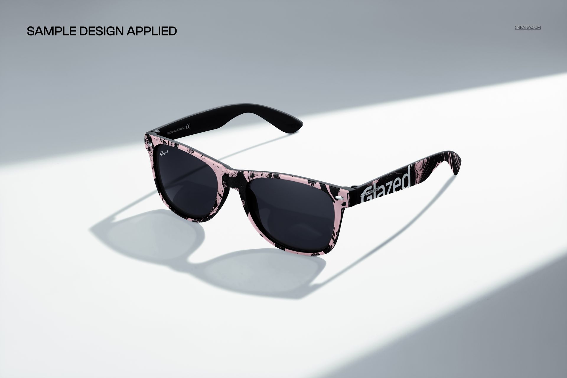 Front angled view of sunglasses mockup with dark lenses and pink marbled pattern on the frames, placed on a sunlit surface.
