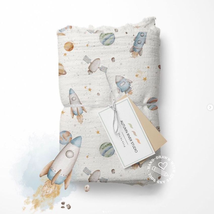 Muslin swaddle blanket mockup featuring a hand-drawn space pattern with rockets, planets, and stars on a white background.